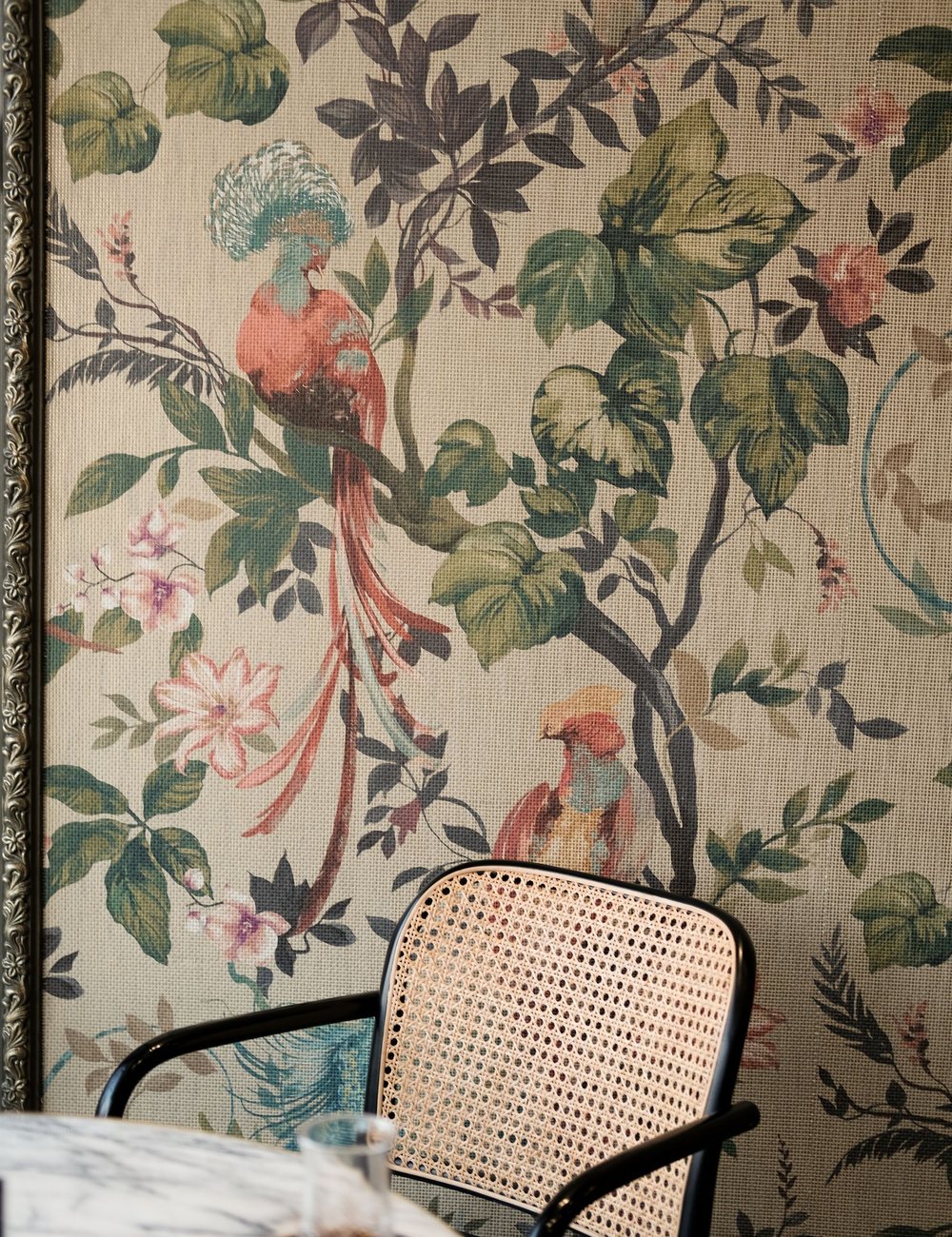 Feature wall in botanical-and-bird chinoiserie wallpaper beside a caned chair